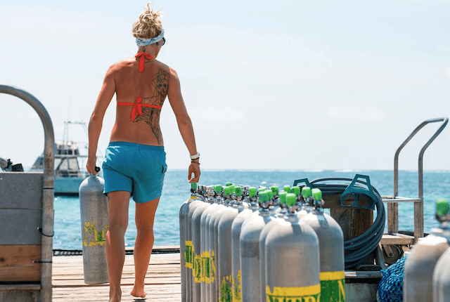 A person in a red bikini top and blue shorts is carrying a scuba tank on a wooden pier. They have a tattoo on their back and are walking away from the camera. Several scuba tanks are lined up nearby, with the ocean and a boat in the background.